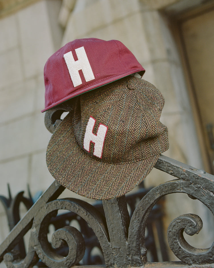 Two Ebbets Field Harvard University Herringbone Ballcaps, featuring a prominent white letter “H” on the front—one in maroon and the other in brown herringbone—are stacked atop a decorative black iron fence, with a blurred stone building in the background.