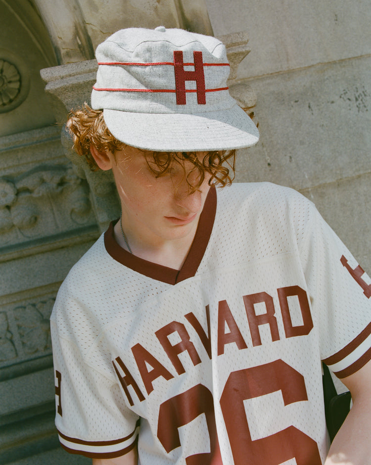 Teen with curly hair wearing an Ebbets Field Harvard University 1899 Pillbox Wool Ballcap and a white Harvard 26 jersey stands outside by a stone wall, looking downward.