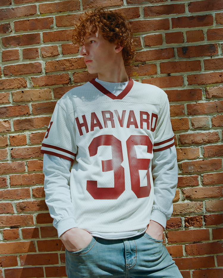 A person with curly hair wearing the Ebbets Field Harvard University Mesh Football Jersey in white, featuring the number 36, pairs it with jeans while standing against a brick wall and looking to the side with hands in their pockets.