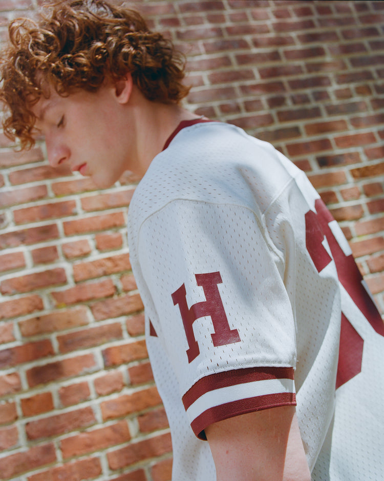 A young person with curly hair is wearing an Ebbets Field Harvard University Mesh Football Jersey, featuring red trim and the letter H, as they stand thoughtfully near a brick wall and look down.