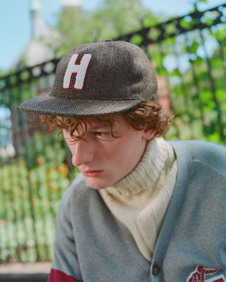 A young person with curly hair sits outdoors by a black metal fence and greenery, wearing the Ebbets Field Harvard University Herringbone Ballcap with a large H on it and a cream turtleneck sweater under a gray jacket.