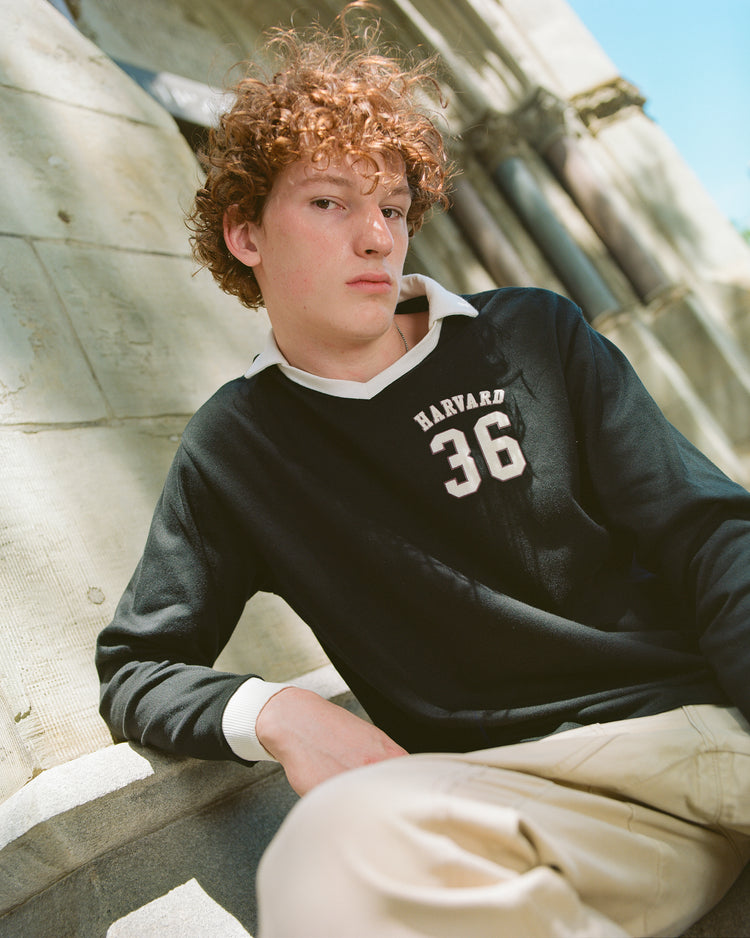 A young person with curly hair wears the black Harvard University Vintage Soccer Goalie Jersey by Ebbets Field and beige pants, sitting outdoors against a stone wall and looking confidently at the camera in natural sunlight.