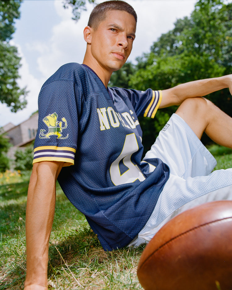 A person wearing the Ebbets Field Notre Dame Mesh Football Jersey in navy and white shorts sits on grass outdoors with a football in front. Trees and a house are visible in the background as the person gazes past the camera.