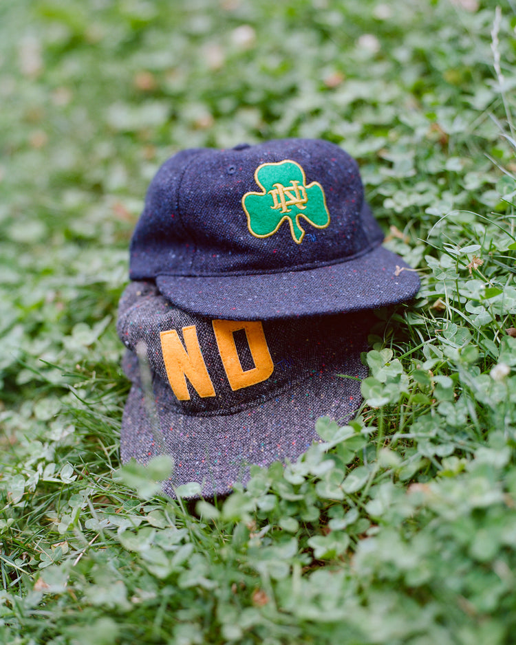 Two dark-colored caps lie on green grass. The top cap, the Ebbets Field Notre Dame X Magee Irish Donegal Wool Ballcap, features a green shamrock and yellow letters; the bottom cap displays large yellow “NO” letters. Both caps are surrounded by clover leaves and small plants.