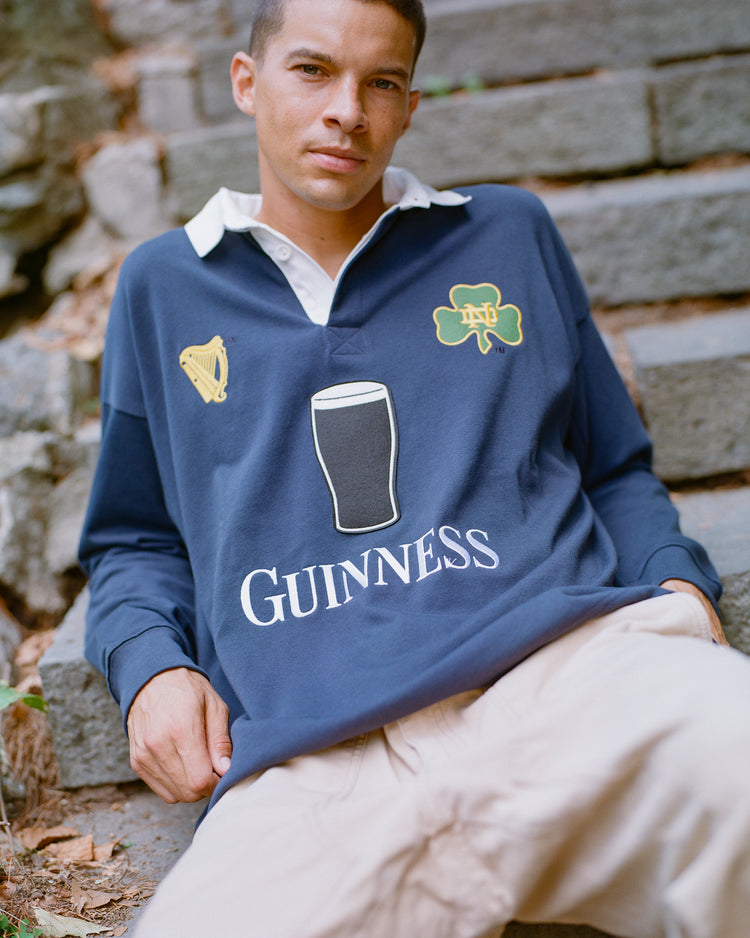 A person sits on stone steps outdoors, wearing the Ebbets Field Notre Dame X Guinness Rugby Shirt—adorned with a harp, shamrock, and pint graphic in navy—celebrating Irish heritage and paired with light pants.