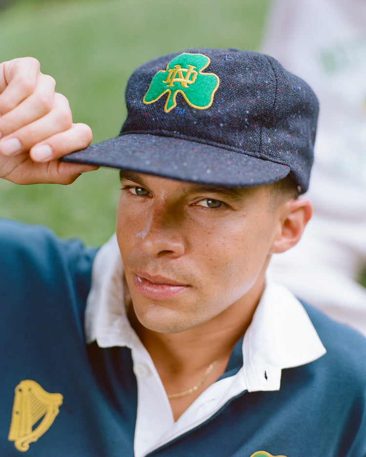Sure! Here’s a rewrite of the sentence using the product data: A person wearing an Ebbets Field Notre Dame X Magee Irish Donegal Wool Ballcap, featuring a green shamrock and ND logo, and dressed in a navy collared shirt with a harp emblem, looks directly at the camera while holding the brim of their cap.