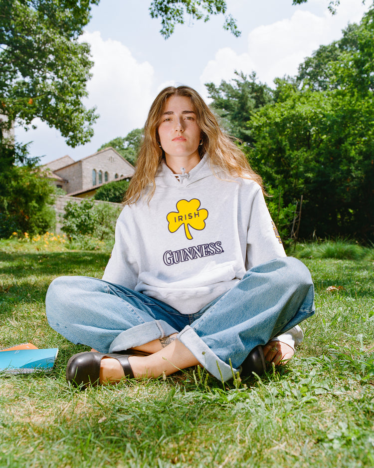 A person with long hair sits cross-legged on the grass, wearing an Ebbets Field Notre Dame X Guinness Heavyweight Pullover Hoodie in light gray, paired with blue jeans and black shoes. Trees and a building are in the background, emphasizing their Irish heritage.
