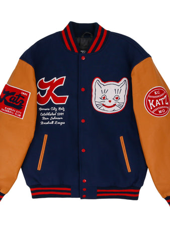 The Kansas City Katz Dna Varsity Jacket by Ebbets Field features navy blue body, tan sleeves, red and white striped cuffs, and embroidered patches including a smiling cat face, logos, and text on the chest and sleeves.