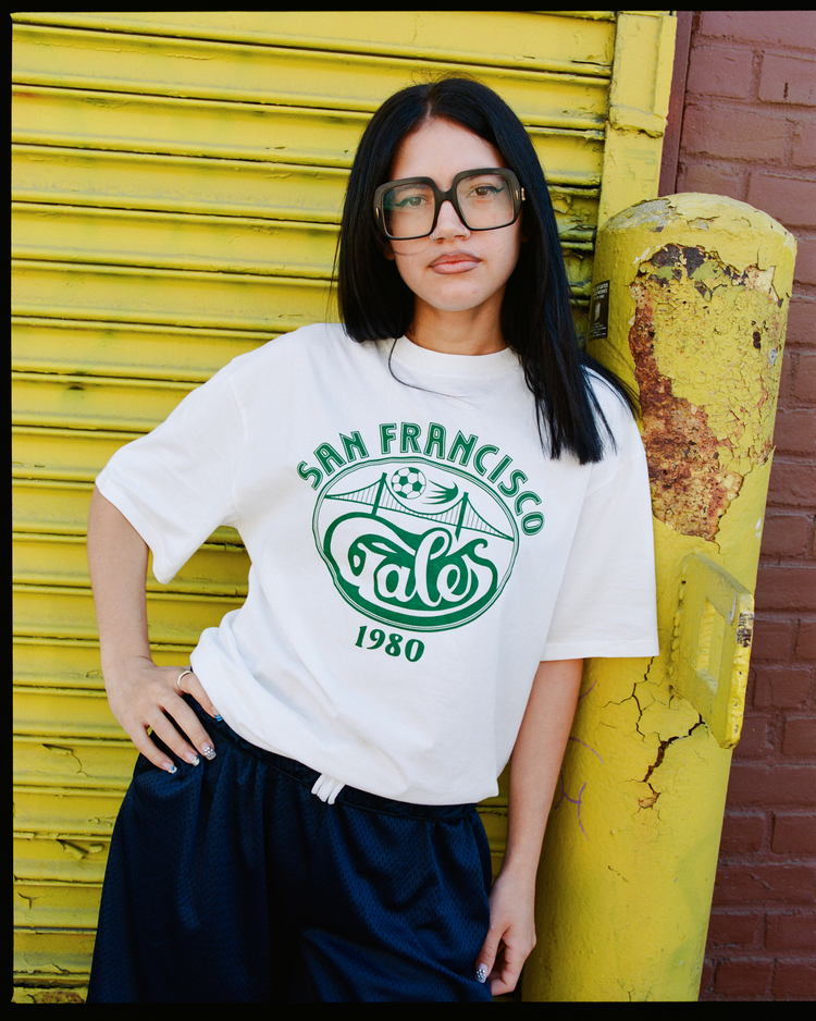 A woman with long dark hair and large glasses wears an Ebbets Field San Francisco Golden Gate Gales Short Sleeve Graphic T-Shirt and dark shorts, standing beside a yellow pole and weathered metal shutters.