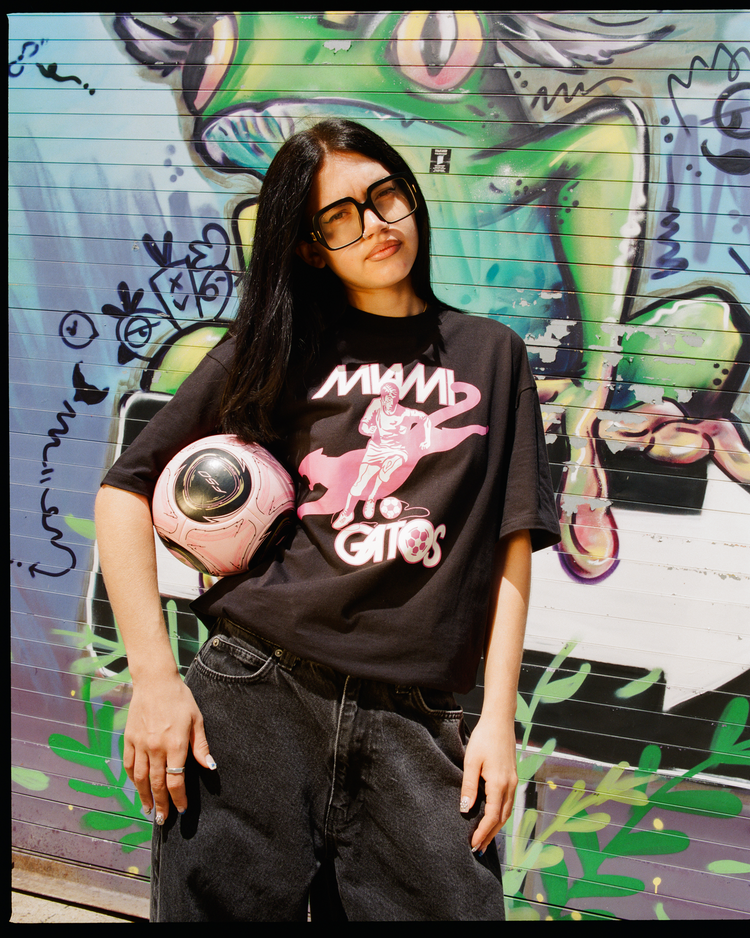 A person with long dark hair and large glasses stands before a colorful graffiti mural, wearing the Ebbets Field Miami Gatos Short Sleeve Graphic T-Shirt and baggy jeans, holding a pink helmet under one arm.