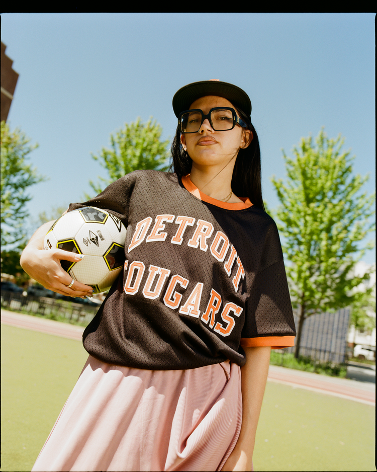A young person in large glasses and a baseball cap holds a soccer ball, wearing the Ebbets Field Detroit Cougars Mesh Soccer Jersey in black with orange lettering, paired with a pink skirt.