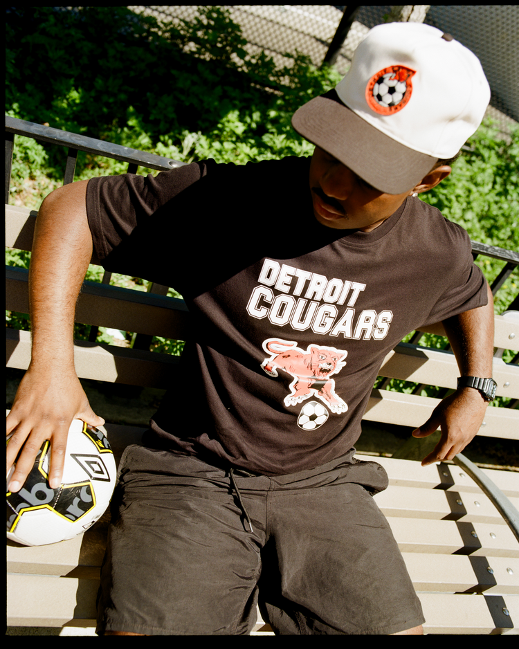 A person sits on a bench outdoors near greenery and a fence, wearing an Ebbets Field Detroit Cougars A-Frame Cotton Twill Ballcap and resting one hand on a soccer ball.