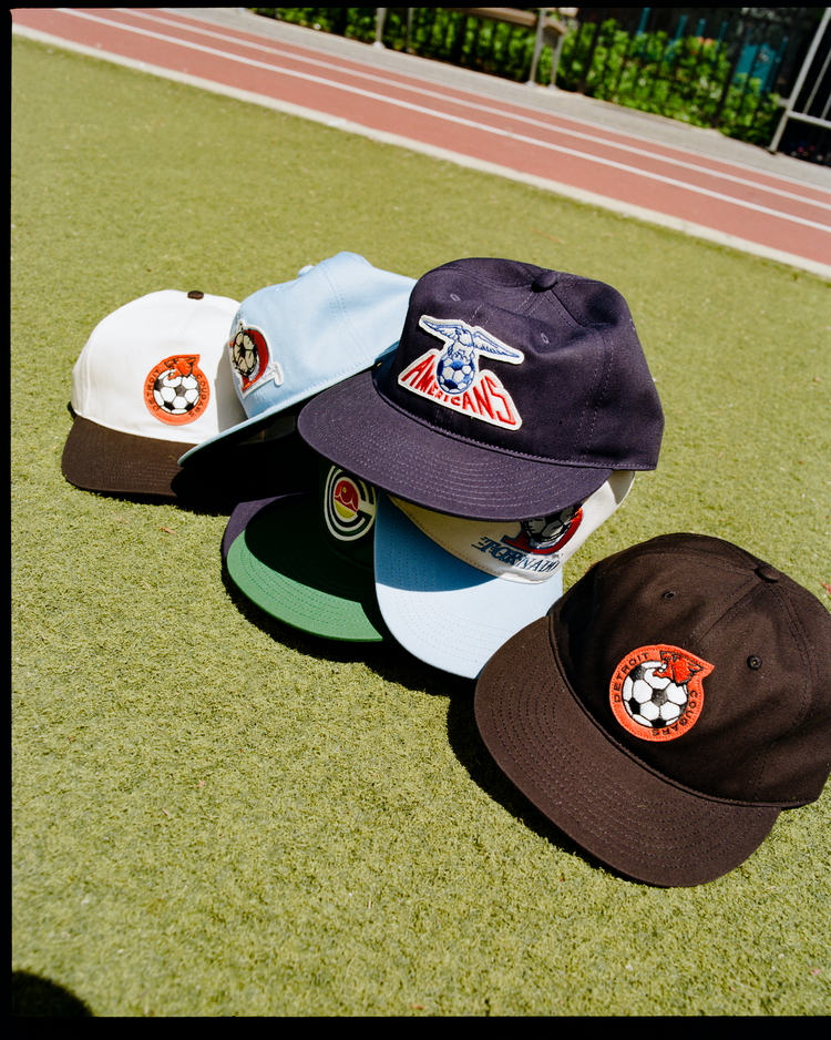 Five Ebbets Field New Jersey Americans Cotton Twill Ballcaps, each with unique soccer-themed patches and colors, are displayed on green turf beside a running track with a fence in the background.