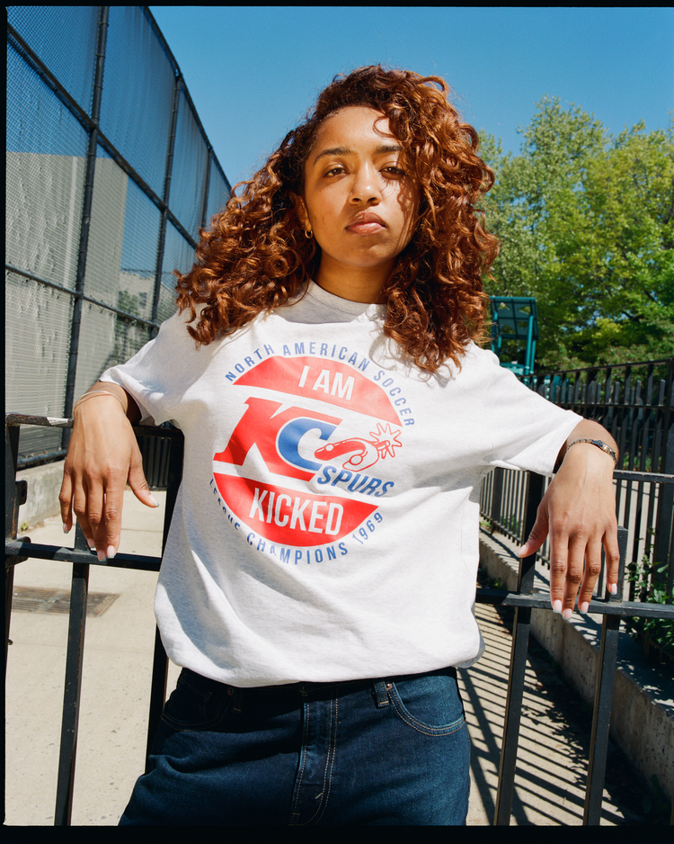 A woman with curly auburn hair leans on a black metal fence, wearing the Ebbets Field Kansas City Spurs Short Sleeve Graphic T-Shirt featuring bold text celebrating their 1969 NASL championship.