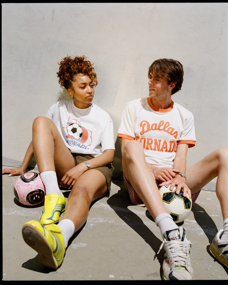 Two people sit on the ground against a light wall, each holding a soccer ball. The person on the right wears the Ebbets Field Dallas Tornados Short Sleeve Graphic T-Shirt, celebrating the NASL original team’s soccer legacy.