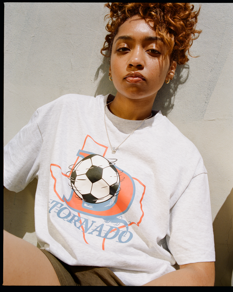 A person with curly hair sits against a light wall, wearing an Ebbets Field Dallas Tornados Short Sleeve Graphic T-Shirt. They have a neutral expression and look directly at the camera.