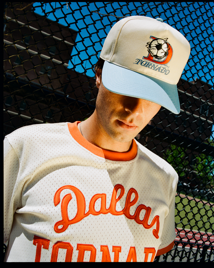 A person wearing a cream Dallas Tornados shirt and the Ebbets Field Dallas Tornados A-Frame Cotton Twill Ballcap stands in front of a chain-link fence on a sunny day, looking down.