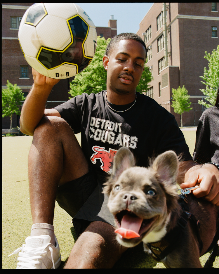 A man wearing the Ebbets Field Detroit Cougars Short Sleeve Graphic T-Shirt sits on a sports field with a soccer ball and a happy gray dog on a leash, apartment buildings and trees in the background.