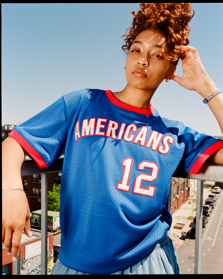 On a balcony with city views and a clear blue sky, a person with curly hair wears the Ebbets Field New Jersey Americans Mesh Soccer Jersey in blue with red trim, celebrating an ASL championship. The jersey features 