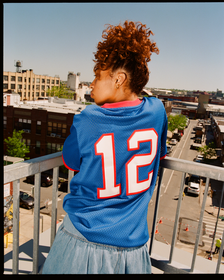 A young woman with curly red hair stands on a balcony, facing away from the camera, wearing an Ebbets Field New Jersey Americans Mesh Soccer Jersey with the number 12 on the back.
