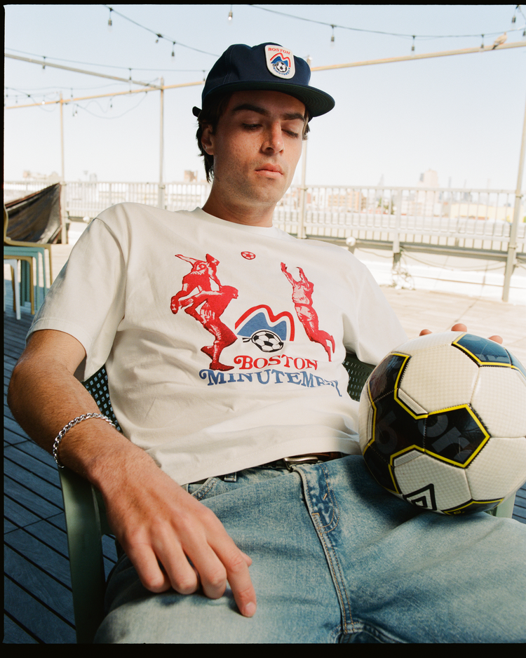 A young man in an Ebbets Field Boston Minutemen Short Sleeve Graphic T-Shirt, blue jeans, and a baseball cap sits outdoors on a deck chair with a soccer ball on his lap. String lights and buildings are visible in the background.