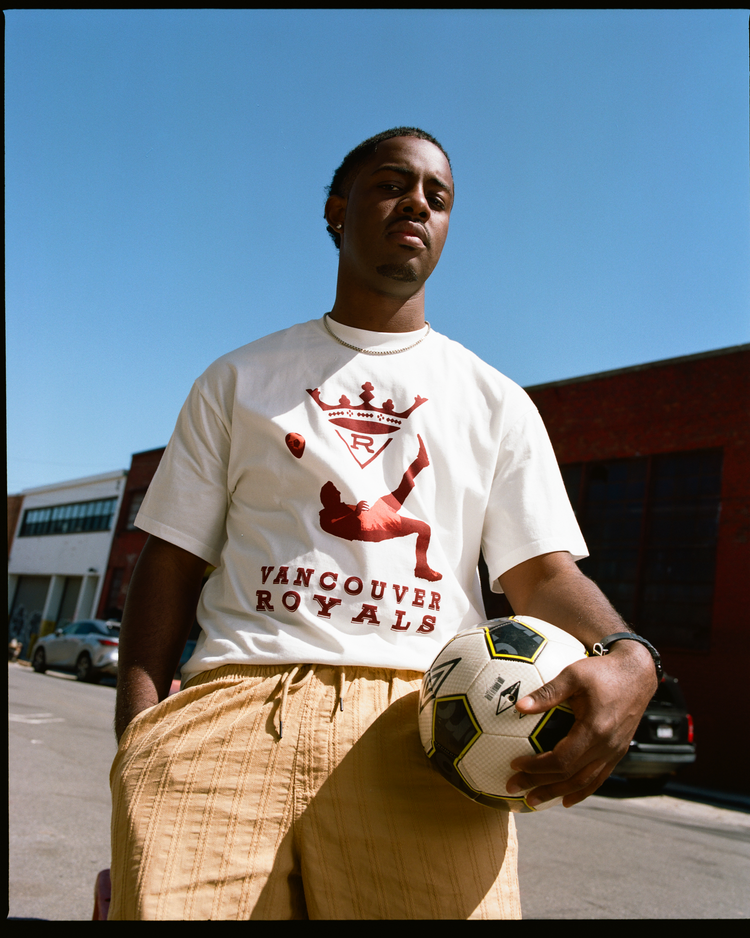 A young man stands outside holding a soccer ball, wearing the Ebbets Field Vancouver Royals Short Sleeve Graphic T-Shirt in white with a red design and tan shorts. The clear blue sky and brick buildings serve as the backdrop.