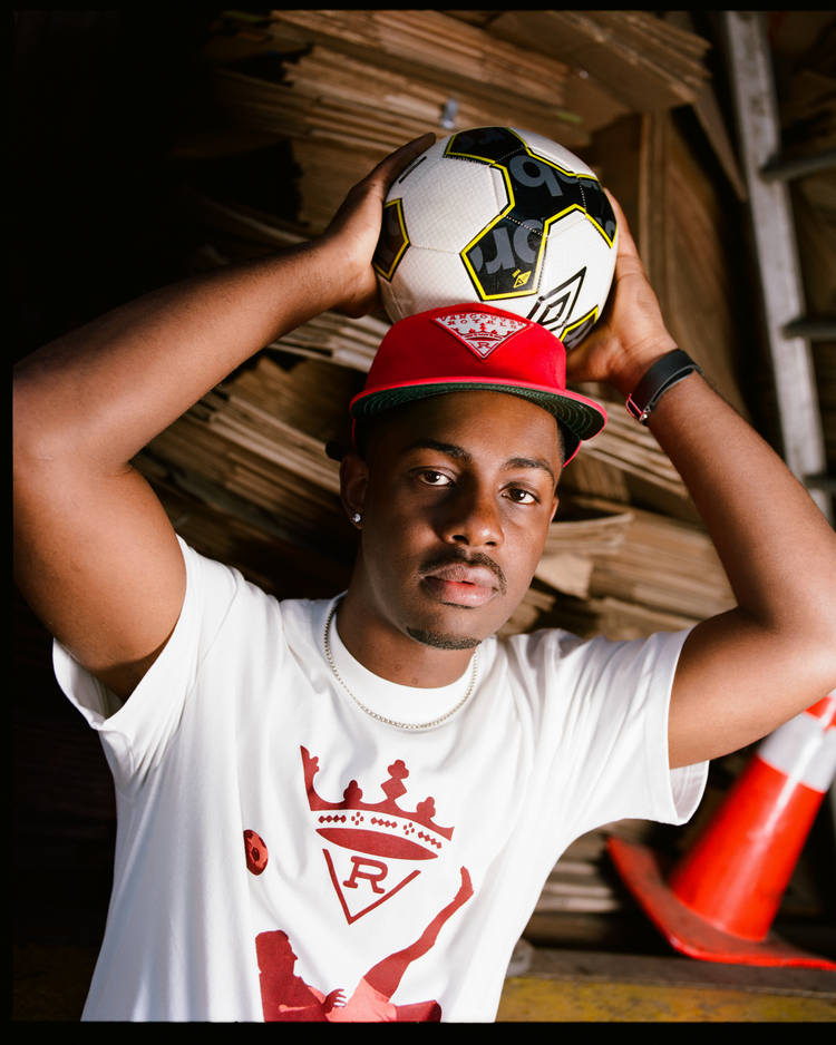 A young man wears an Ebbets Field Vancouver Royals Cotton Twill Ballcap and a white graphic t-shirt, holding a soccer ball above his head. He stands before stacked cardboard and an orange cone, gazing confidently at the camera.