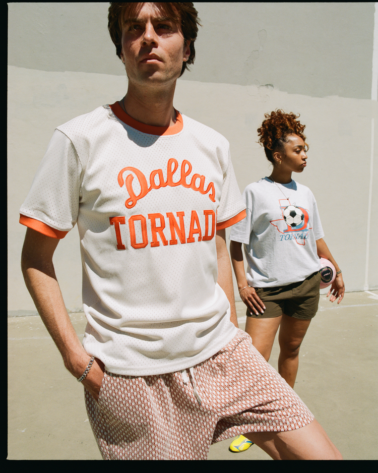 A man in an Ebbets Field Dallas Tornado Mesh Soccer Jersey stands by a woman in a gray shirt with a Texas outline and soccer ball, both posing in front of a wall.