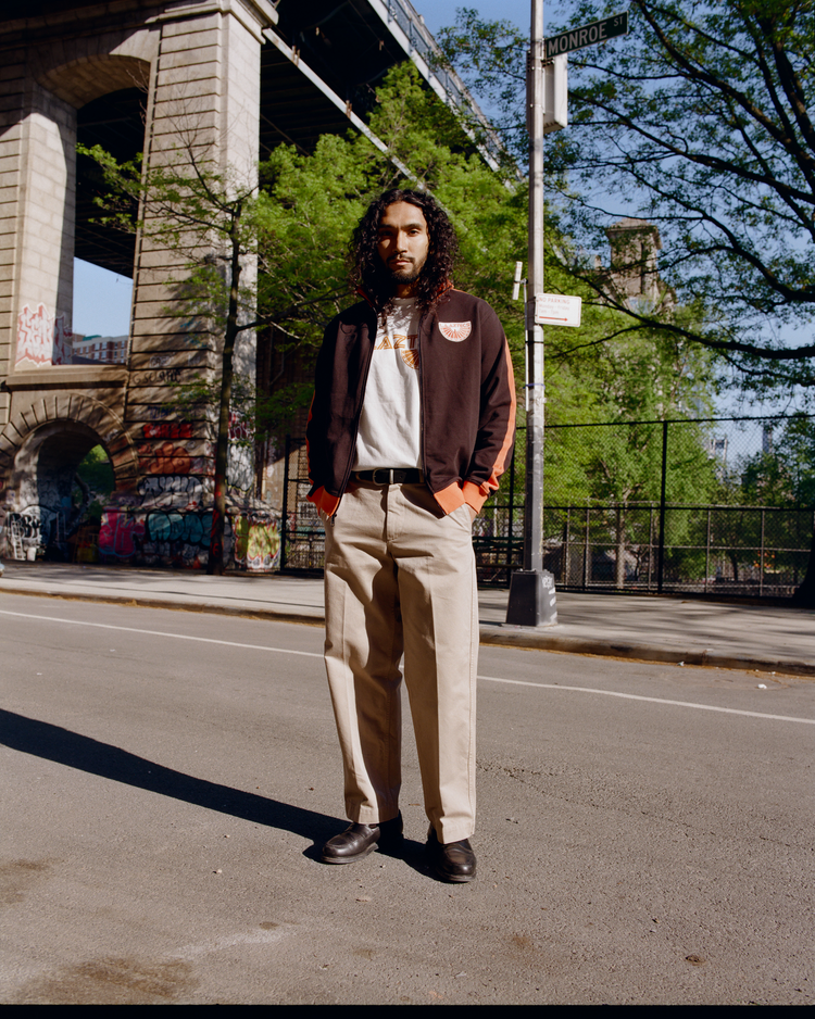 A person with long curly hair stands on a city street near a bridge, wearing an Ebbets Field Los Angeles Aztecs Soccer Warm Up Jacket over a white t-shirt, beige pants, and black shoes. Trees, graffiti, and a Monroe St. sign are in the background.