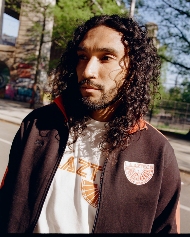 A bearded person with long curly hair wears an Ebbets Field Los Angeles Aztecs Soccer Warm Up Jacket in black and orange over a white AZTECS tee while standing by an outdoor basketball court with trees and graffiti in the background.