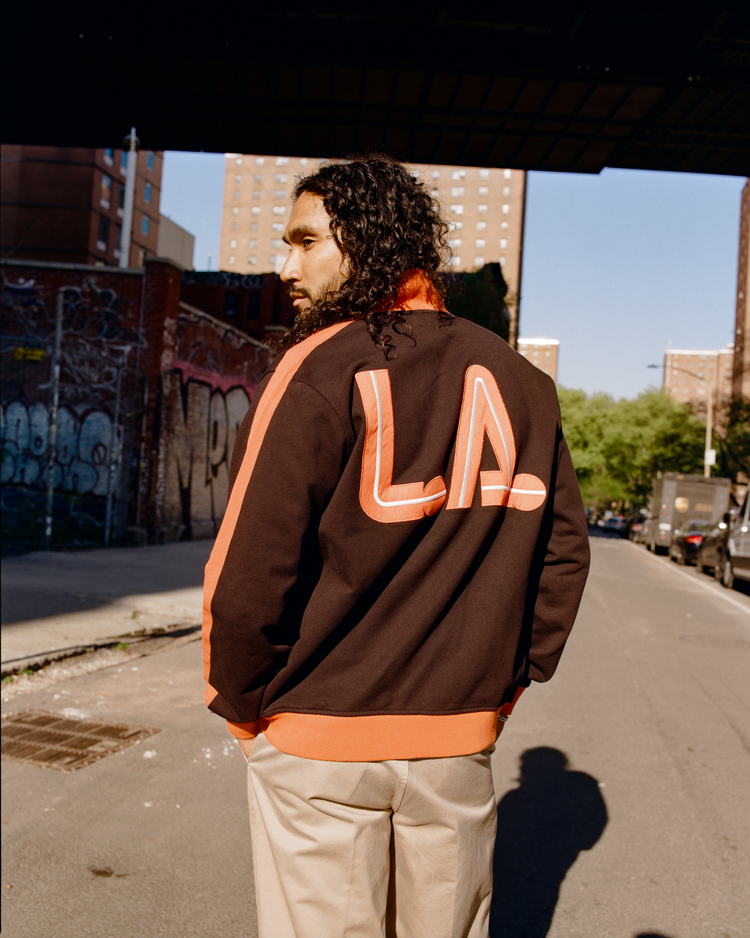 A man with long curly hair stands on a city street, facing away, wearing an Ebbets Field Los Angeles Aztecs Soccer Warm Up Jacket. Apartment buildings and graffiti-covered walls are visible in the background.