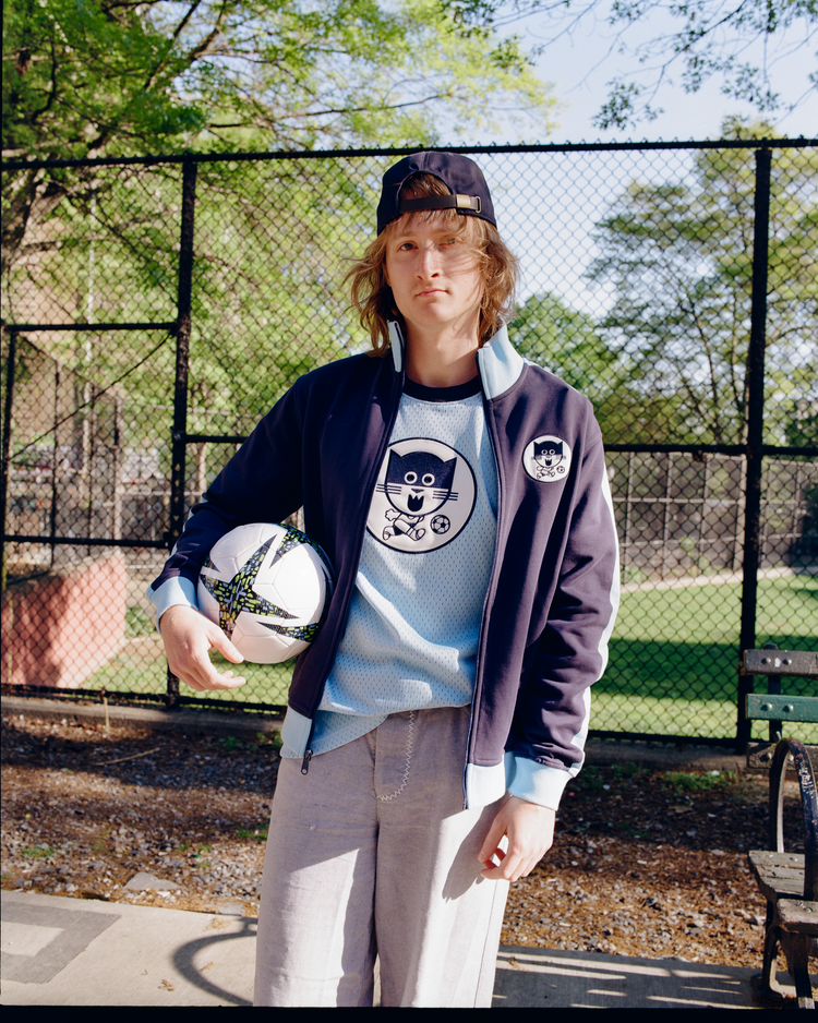 Wearing the Ebbets Field Chicago Cats Soccer Warm Up Jacket and light pants, a long-haired person in a backwards cap stands outdoors holding a soccer ball, with a fence, bench, and green grass visible in the background.