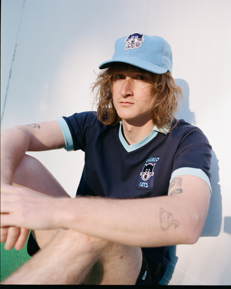 A person with long hair, arm tattoos, and a navy cat-logo T-shirt sits against a white wall wearing the Ebbets Field Chicago Cats Wool Ballcap, looking slightly away from the camera.