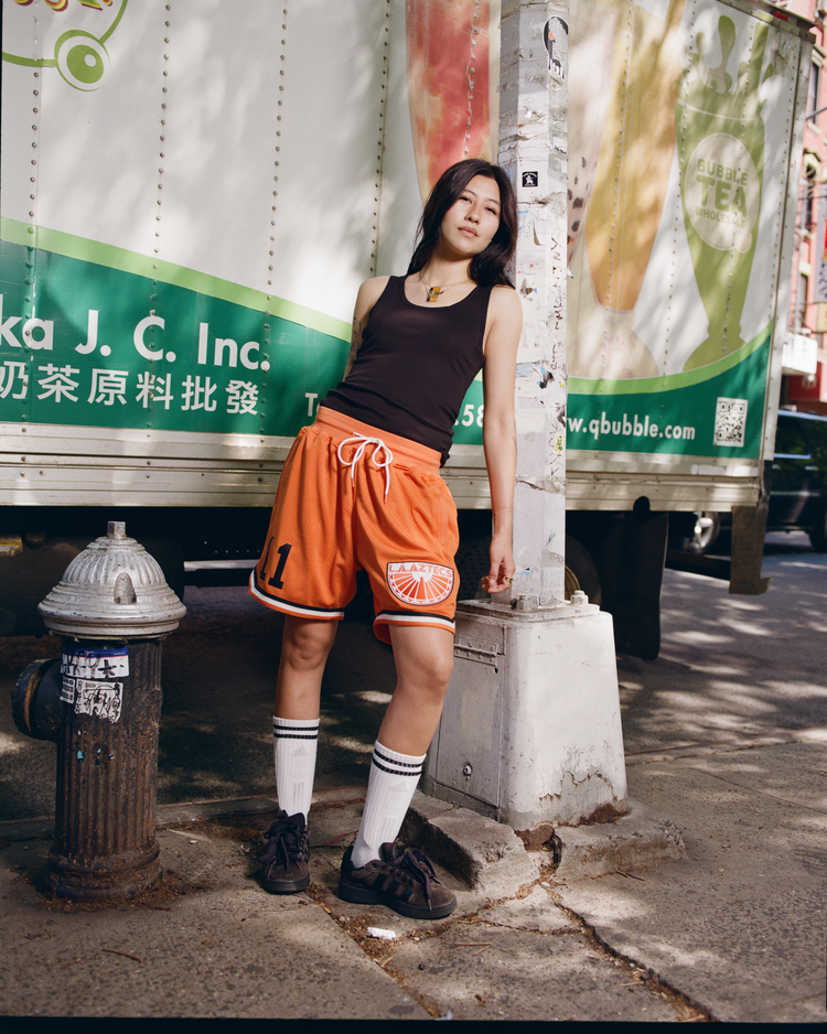 Wearing Ebbets Field Los Angeles Aztecs Mesh Soccer Shorts, a person in orange mesh shorts and a black tank top leans on a city utility pole beside a green-and-white delivery truck with Chinese and English text.