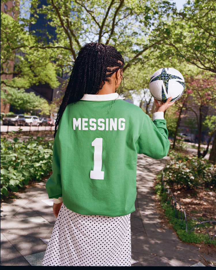 A person with long braids, seen from behind, wears an Ebbets Field New York Cosmos Vintage Soccer Goalie Jersey (number 1). They stand on a park path holding a soccer ball, with trees and buildings in the background.