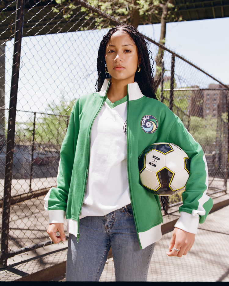 A woman with long braids stands outdoors by a chain-link fence, wearing an Ebbets Field New York Cosmos Soccer Warm Up Jacket and jeans, holding a soccer ball under her arm. Trees and buildings are visible in the background.