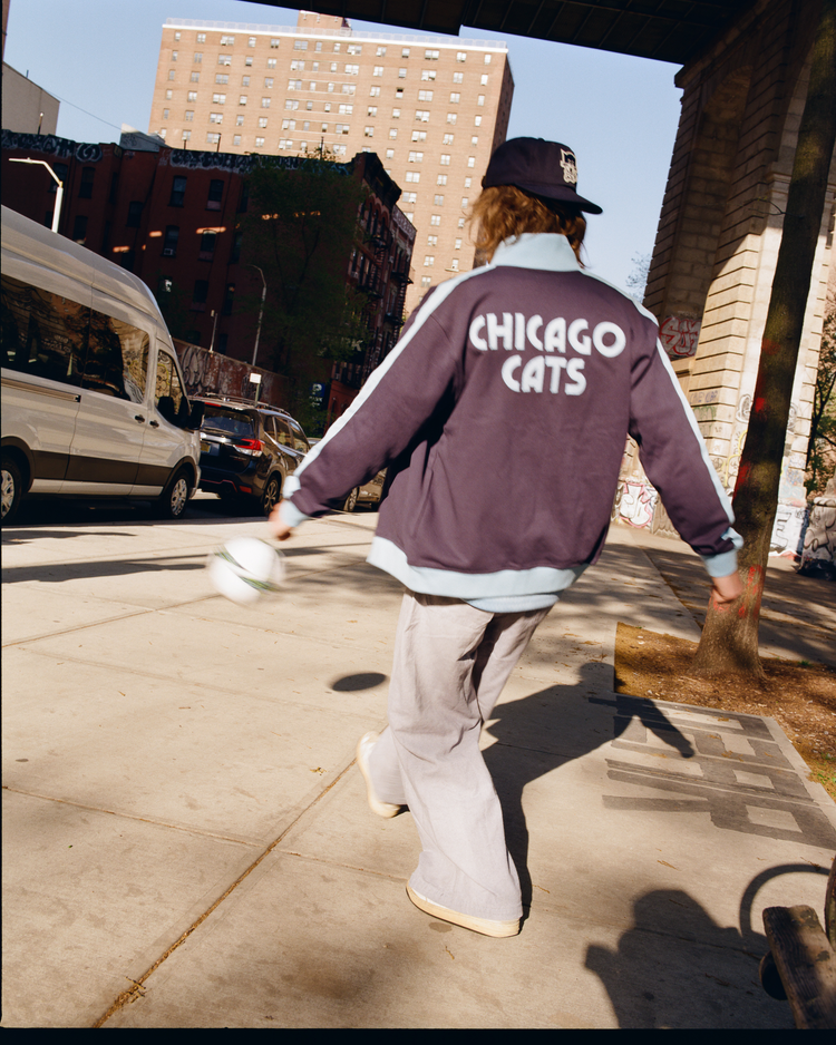 Wearing an Ebbets Field Chicago Cats Soccer Warm Up Jacket, inspired by the American Soccer League, a person plays soccer on a city sidewalk.