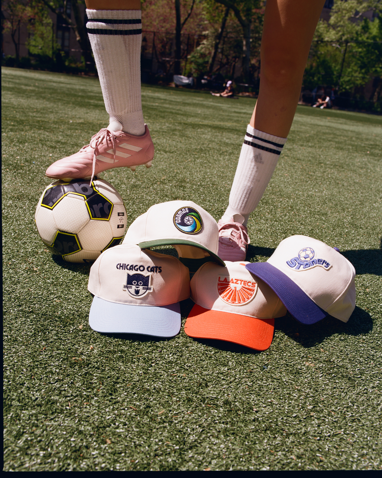 A person in pink sneakers and striped knee-high socks stands on grass with a soccer ball, while four baseball caps—including the Ebbets Field Los Angeles Aztecs A-Frame Cotton Twill Ballcap—are lined up in front.