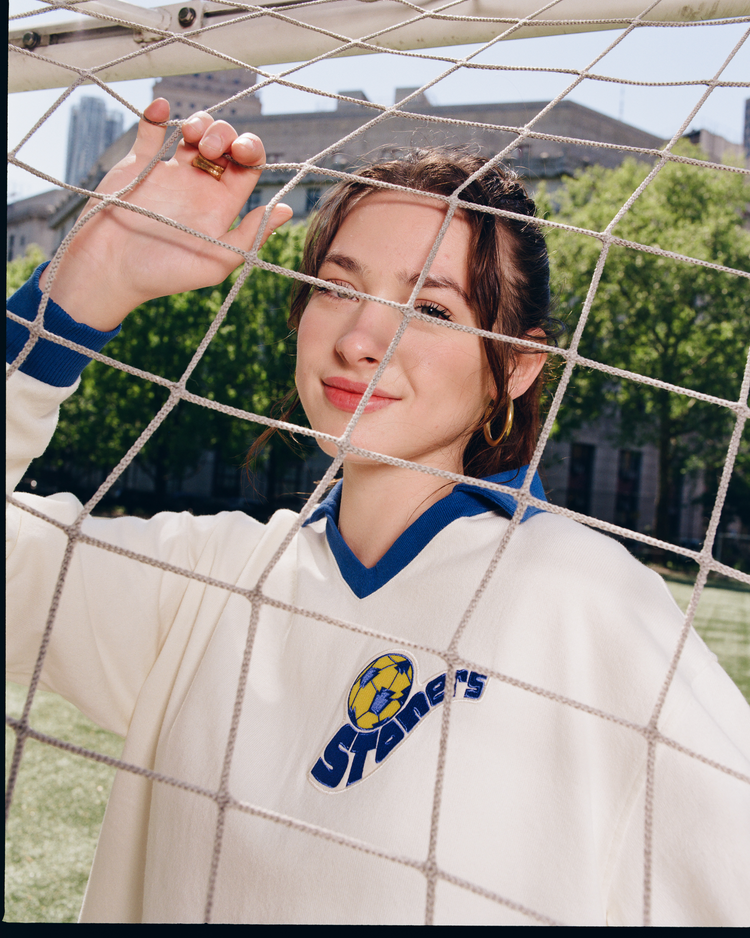 A young woman stands behind a soccer goal net, smiling slightly, wearing the Ebbets Field Pennsylvania Stoner Vintage Soccer Goalie Jersey—a classic white long sleeve with blue trim and a soccer ball logo. Green trees and city buildings are in the background.