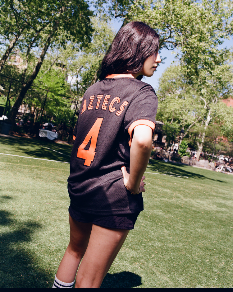 On a grassy field, a person with dark hair wears the Ebbets Field Los Angeles Aztecs Mesh Soccer Jersey, featuring black mesh, orange trim, and “AZTECS” with number 4 on the back. Trees and sunlight fill the background.