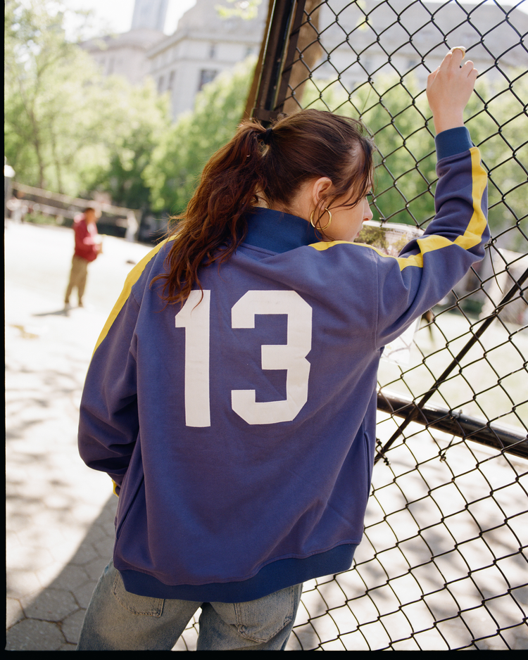 A person with long brown hair in a ponytail leans against a chain-link fence, wearing an Ebbets Field Pennsylvania Stoners Soccer Warm Up Jacket. The blue jacket features yellow stripes and the number 13 on the back. Trees and a building are in the background.