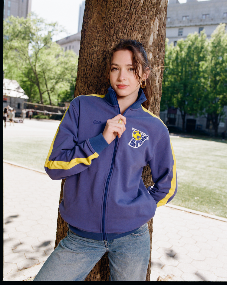 A woman in an Ebbets Field Pennsylvania Stoners Soccer Warm Up Jacket leans against a tree in the park, hands in her pockets and a slight smile on her face, showing off the sporty zip jacket with green grass and tall trees behind her.