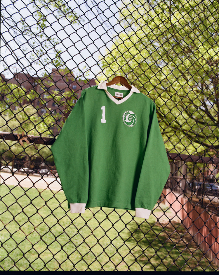 The Ebbets Field New York Cosmos Vintage Soccer Goalie Jersey, featuring a green design, white collar, spiral logo, and number 1, hangs on a wire fence outdoors with trees and grassy field in the background.