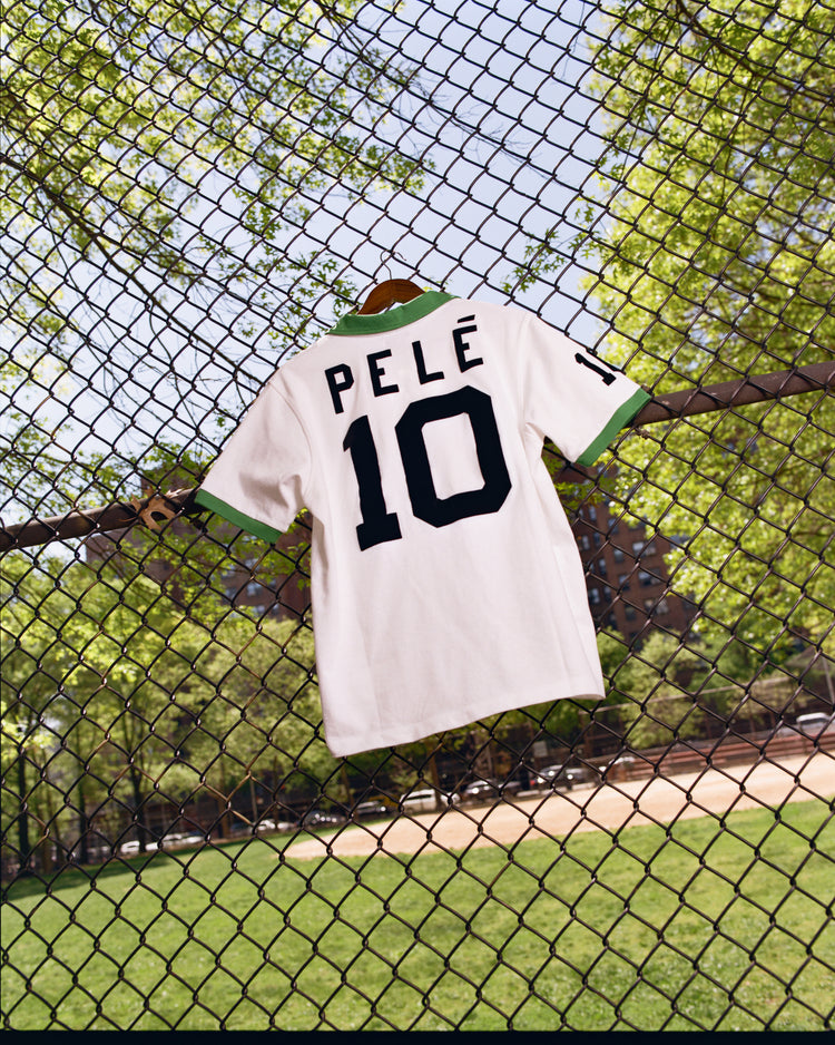 An Ebbets Field New York Cosmos Vintage Collar Soccer Jersey hangs on a fence, its white design and number 10 popping against a sunny park with green trees in the background.