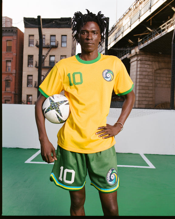 A soccer player in a yellow and green Ebbets Field New York Cosmos Pelé Mesh Soccer Jersey stands on an outdoor court, holding a soccer ball. Buildings and a bridge are in the background, with the number 10 on both jersey and shorts.
