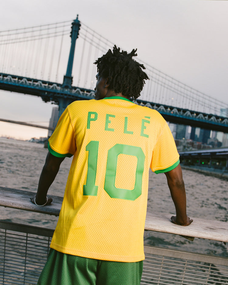 A person in a yellow Ebbets Field New York Cosmos Soccer Mesh Jersey stands by a waterfront railing, looking at a suspension bridge with city buildings in the background, evoking classic Cosmos soccer moments.