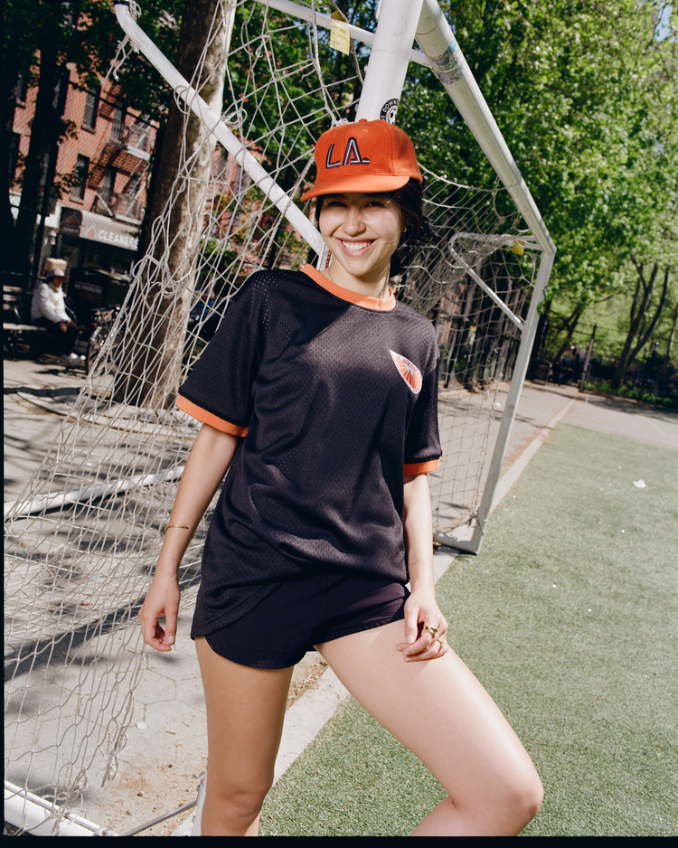 A person in a black sports jersey and shorts smiles near a soccer goal, wearing an Ebbets Field Los Angeles Aztecs Vintage Wool Ballcap, with trees and buildings seen behind on the outdoor field.