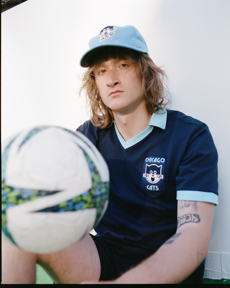 Wearing the Ebbets Field Chicago Cats Vintage Collar Soccer Jersey, a long-haired individual pairs it with a matching cap, sitting before a white wall with a soccer ball—an ideal look for American Soccer League fans.