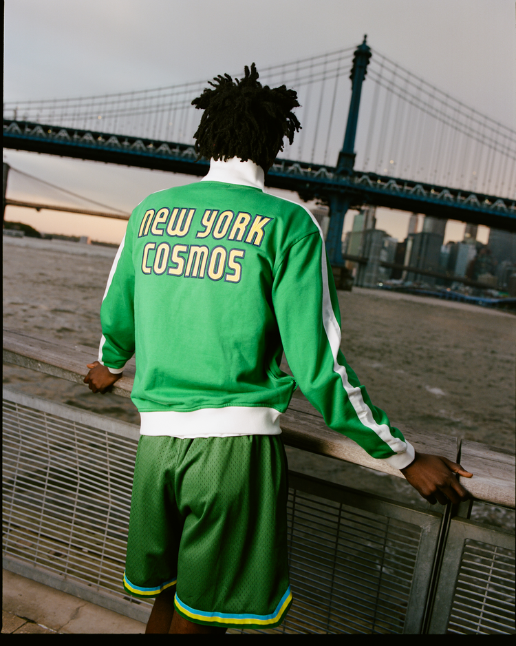 A person in an Ebbets Field New York Cosmos Soccer Warm Up Jacket stands by a river railing, with a bridge and city skyline in the background.