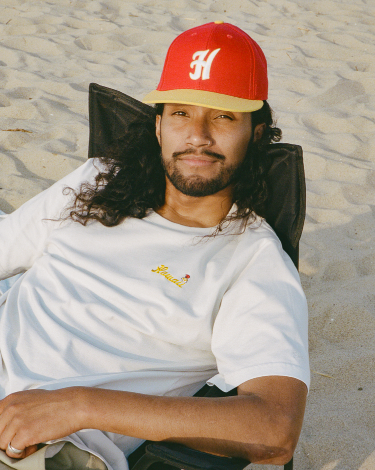 A man with long hair and a beard relaxes on a sandy beach, wearing a white t-shirt and the EFF Hawaii Islanders 1986 Authentic Wool Ballcap.